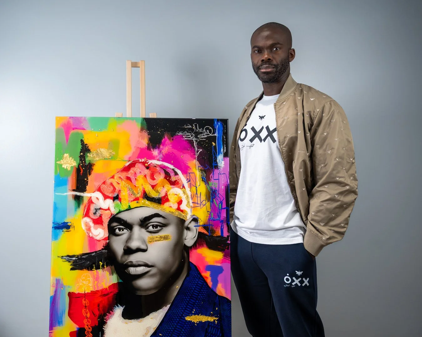 A man standing next to a colorful abstract portrait of a woman with a serious expression, wearing a headscarf. The portrait features bright splashes of colors like pink, yellow, green, and blue, and is displayed on an easel.