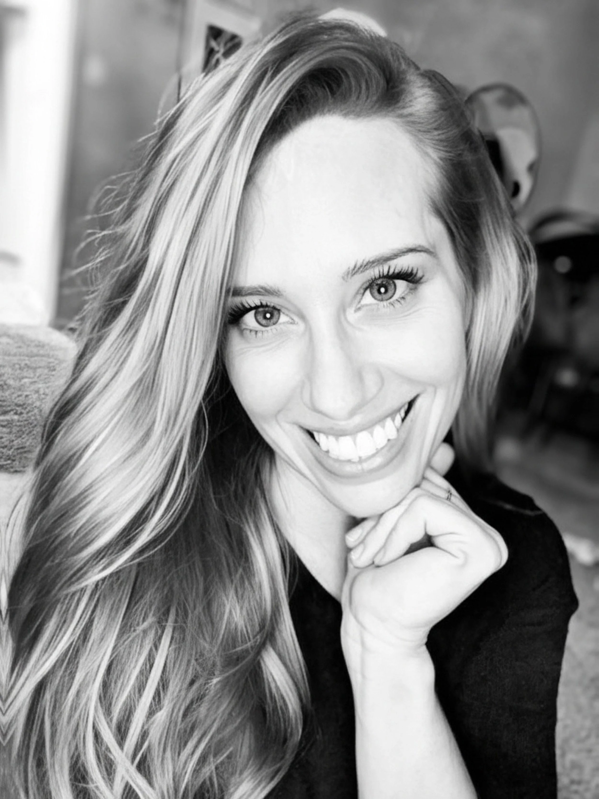 Black and white photo of a woman with long wavy hair smiling, resting her chin on her hand.