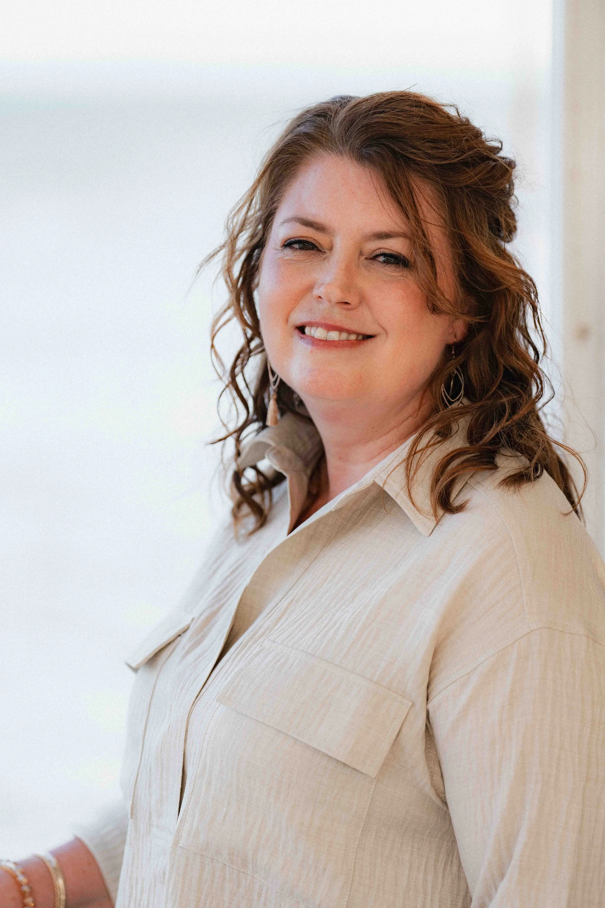 A woman with wavy brown hair smiling, wearing a light-colored collared shirt, and earrings, standing against a white background.