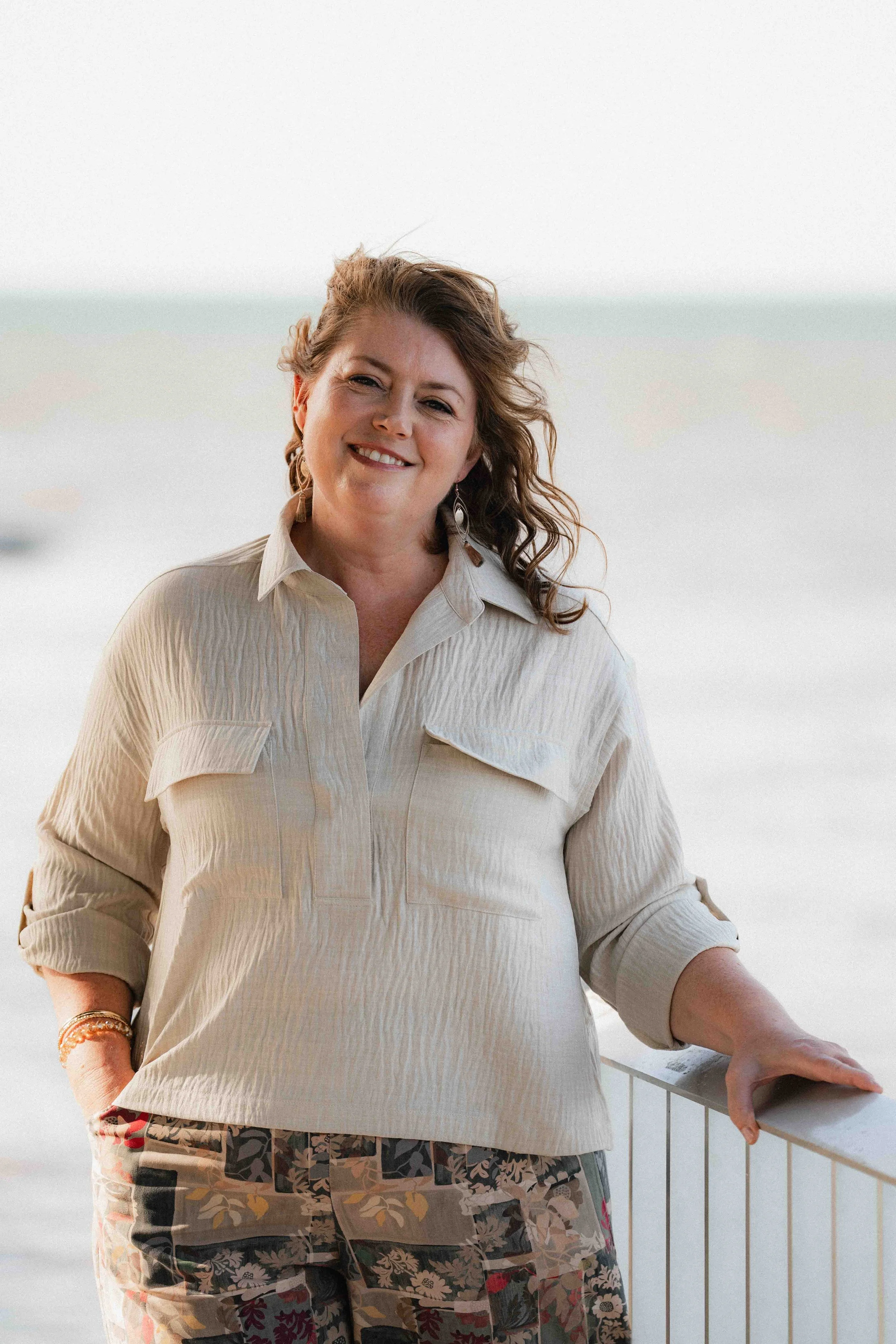 A woman with curly brown hair smiling at the camera, standing outdoors near the beach, wearing a beige shirt and patterned pants.