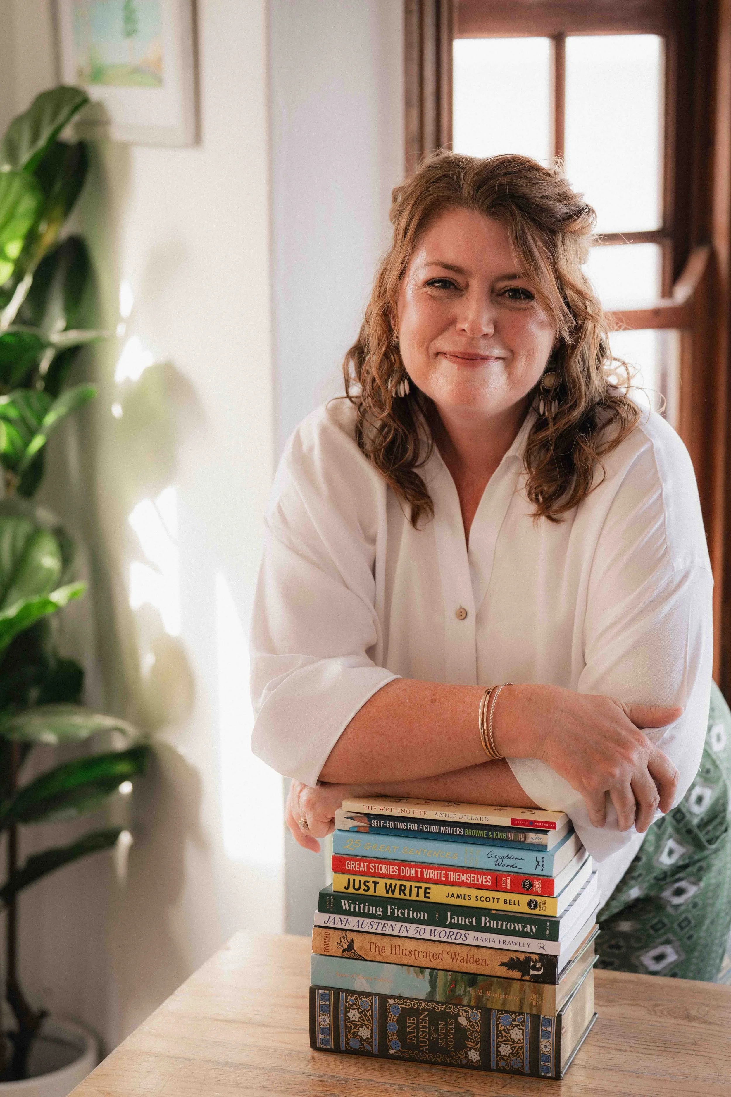 A woman with curly brown hair smiling while leaning on a stack of books on a wooden table, in a room with a large window and a plant.