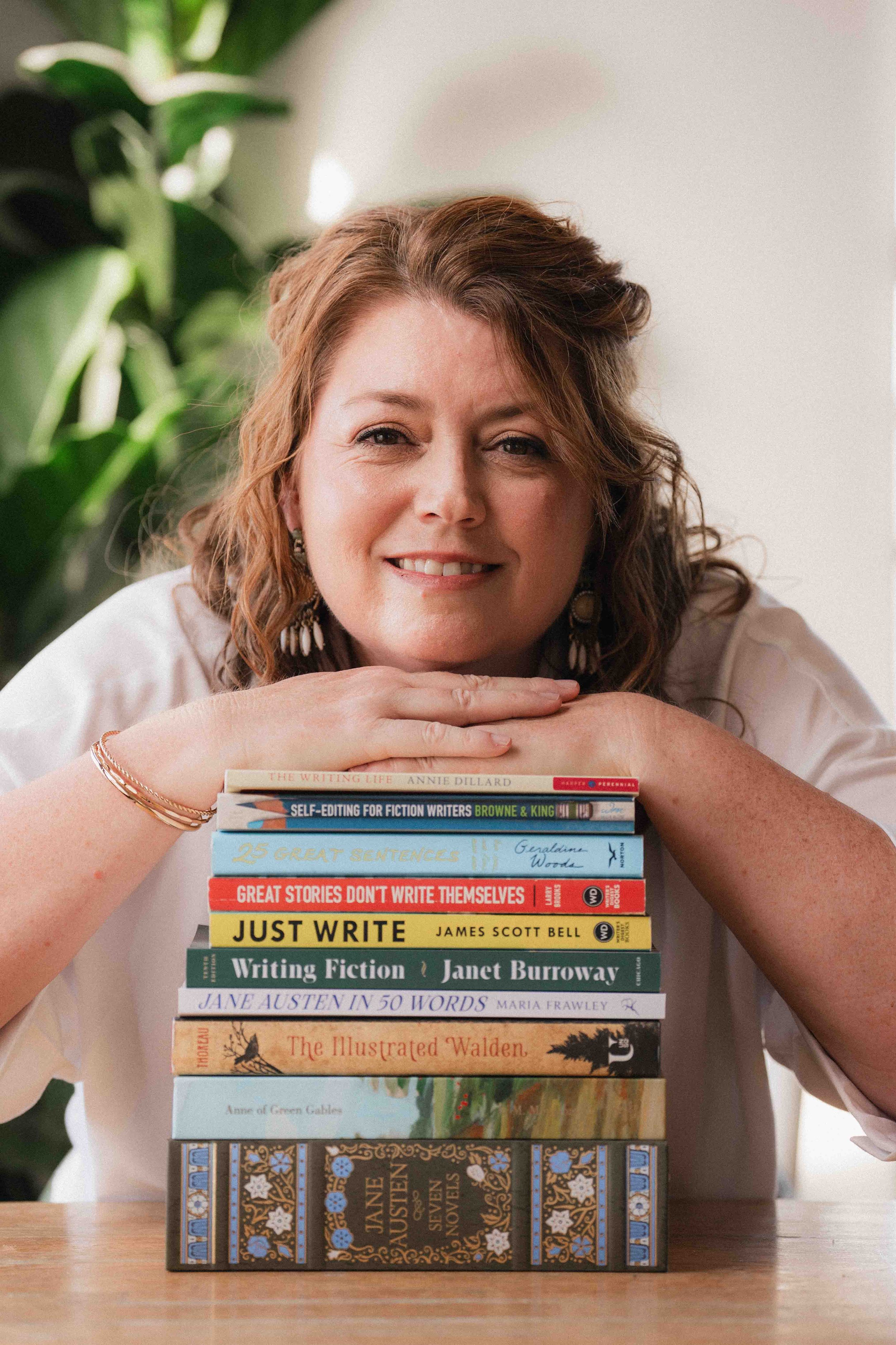 A woman with wavy, reddish-brown hair is leaning on a stack of books and smiling at the camera. There is a large green plant in the background.