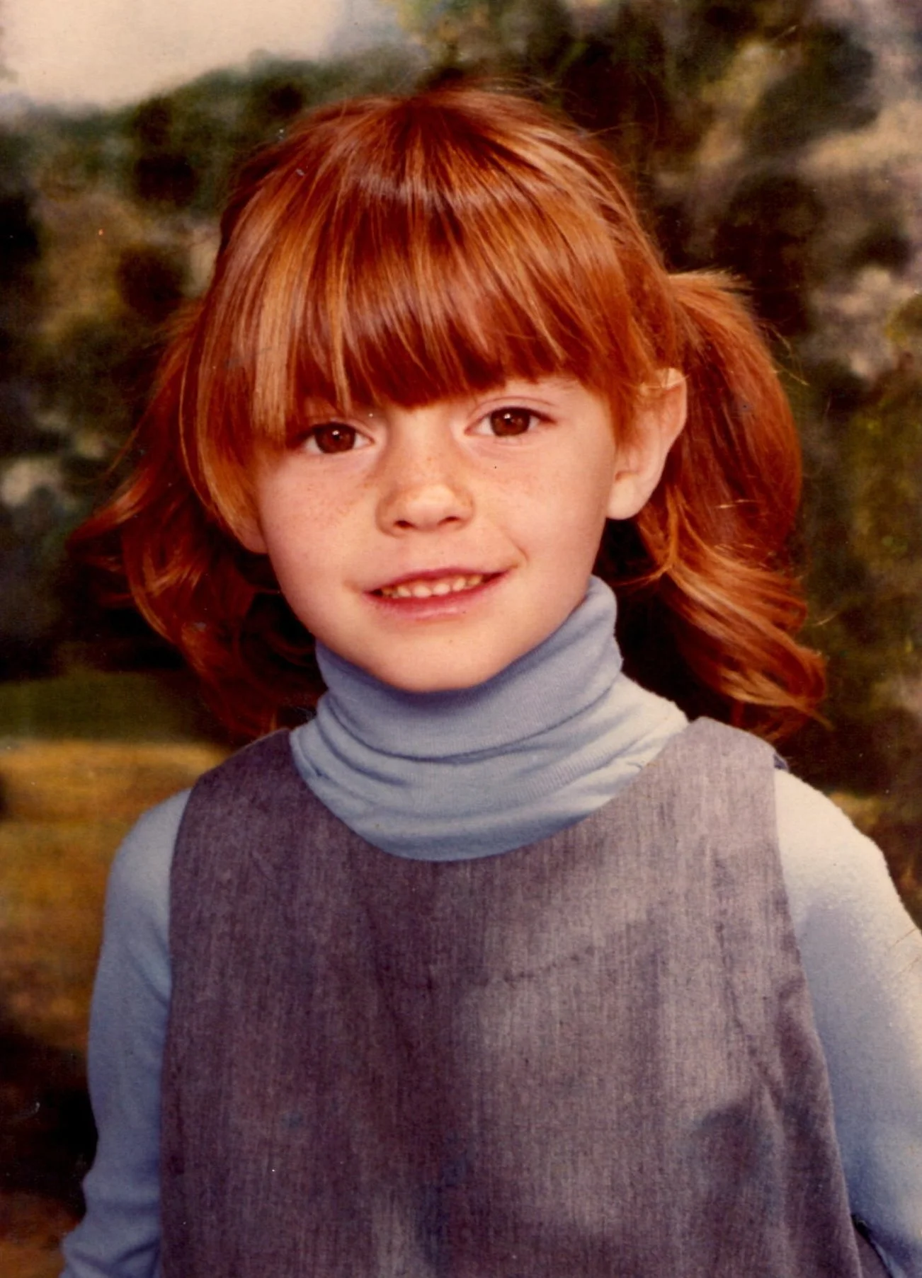 A young girl with red hair styled in pigtails, wearing a gray turtleneck and sleeveless gray dress, smiling outdoors with trees in the background.