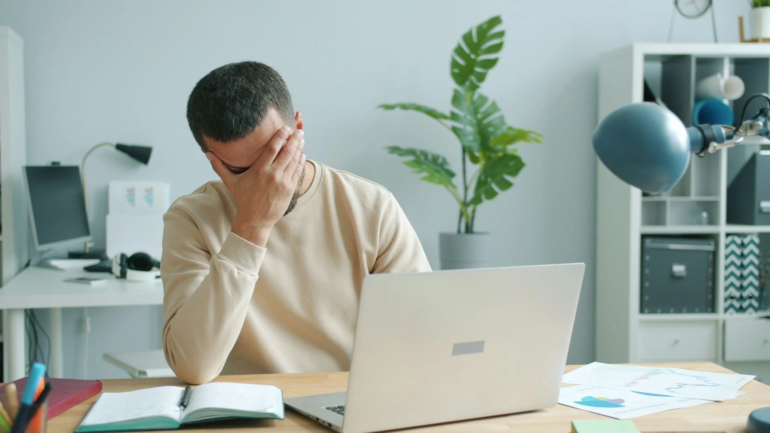 man in cream colored shirt sitting at a desk in front of a silver laptop with his hand covering his face looking dismayed
