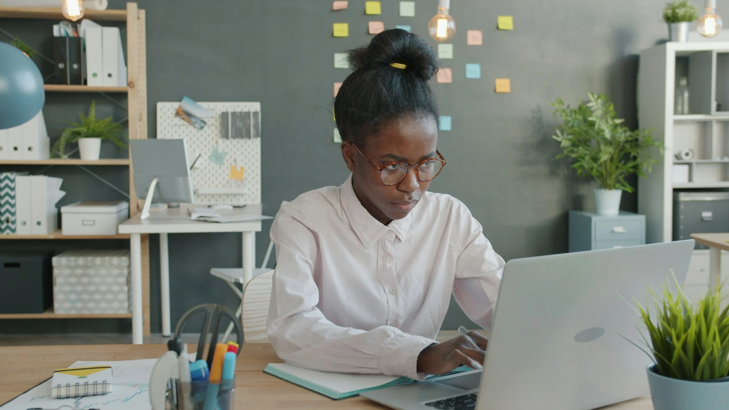 person wearing a white button down shirt and glasses sitting at a desk typing on their silver laptop