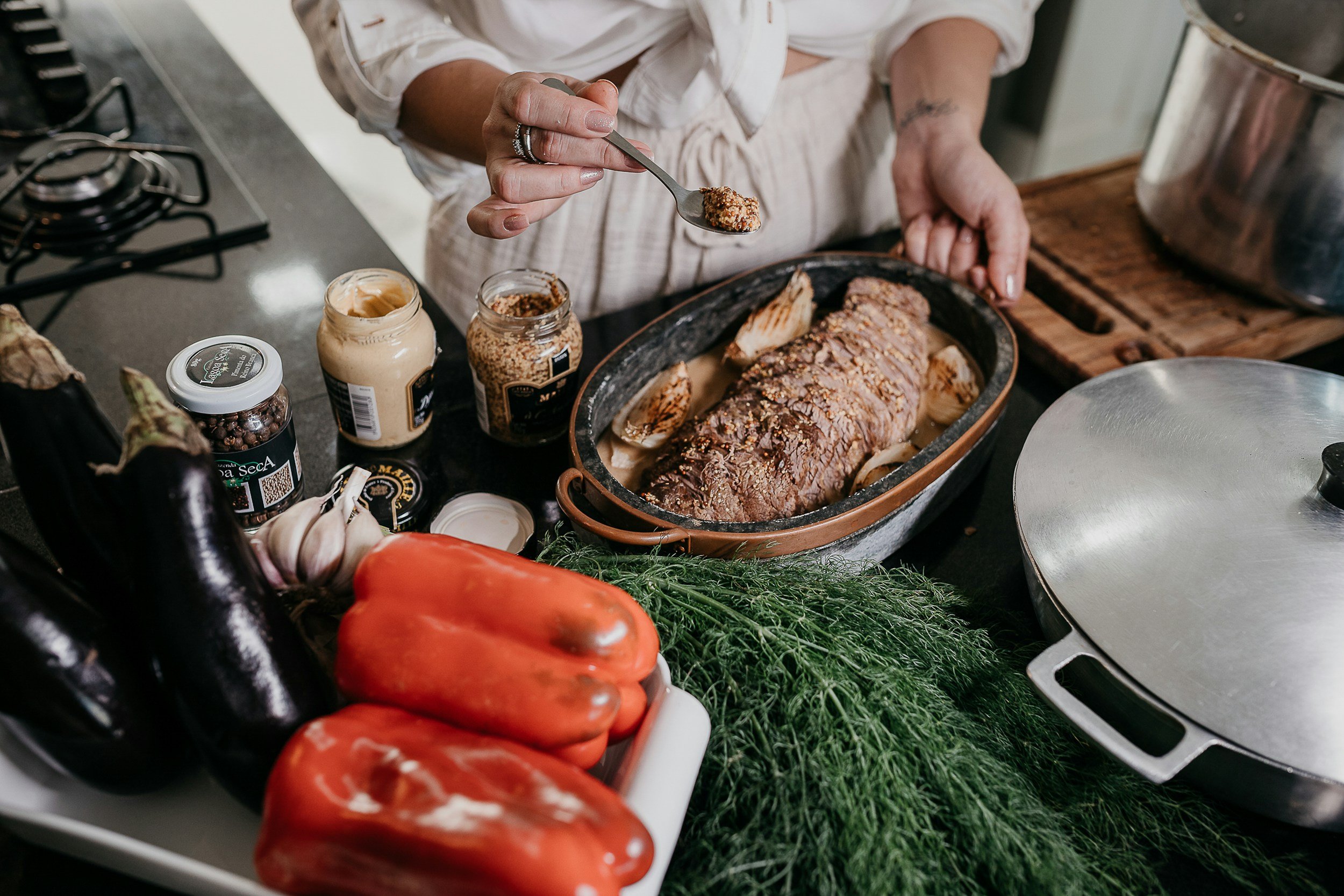 woman in cream colored outfit spooning course ground mustard over a roast surrounded by fresh vegetables and ingredients