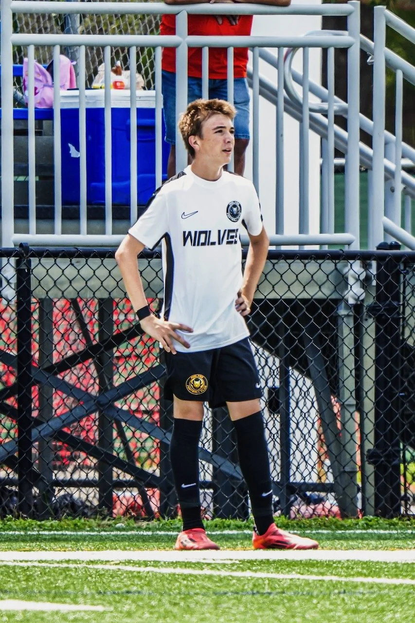 A young male soccer player standing on a field during a game, wearing a white uniform with black shorts, black socks, and red cleats. He has light brown hair and appears to be focused on the game.