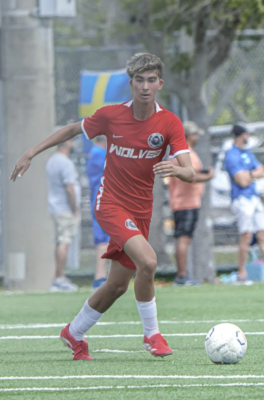 A young male soccer player in a red uniform running on a soccer field with a soccer ball at his feet, with spectators in the background.