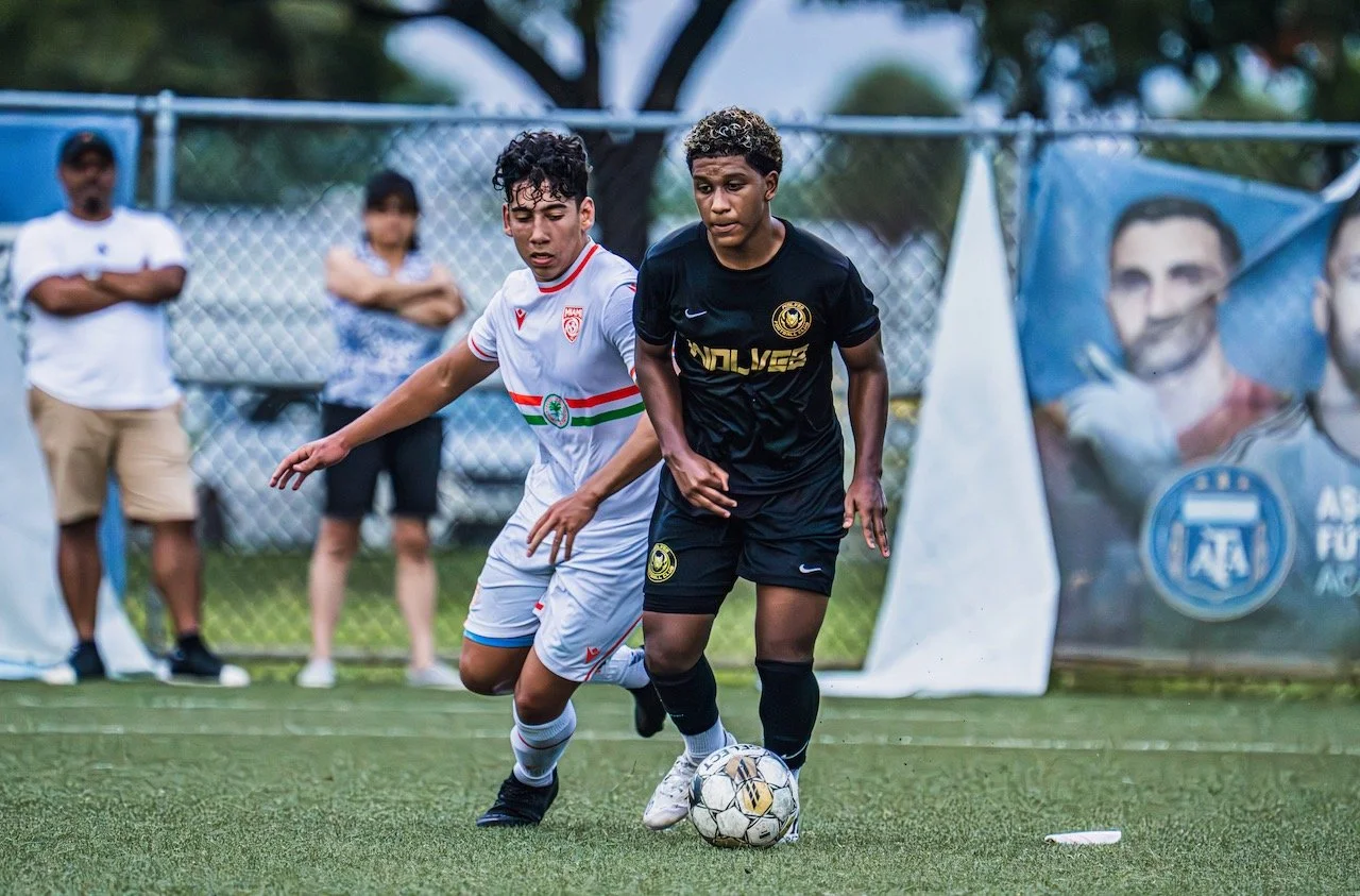 Two young soccer players competing for the ball on the field during a match, with onlookers in the background and a large banner featuring a person’s face.