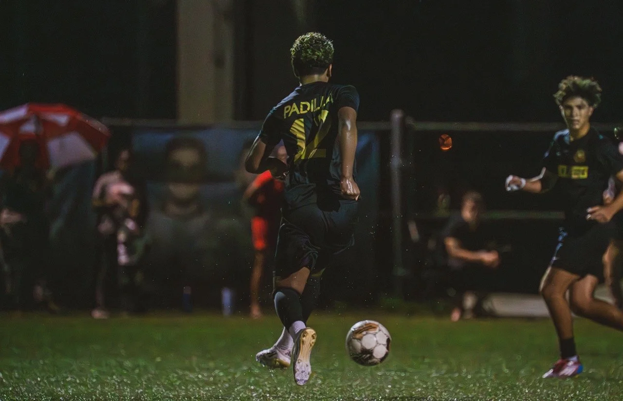 A soccer match taking place at night, with players in black uniforms, one of whom is kicking a soccer ball on a wet field, and spectators watching under umbrellas in the background.