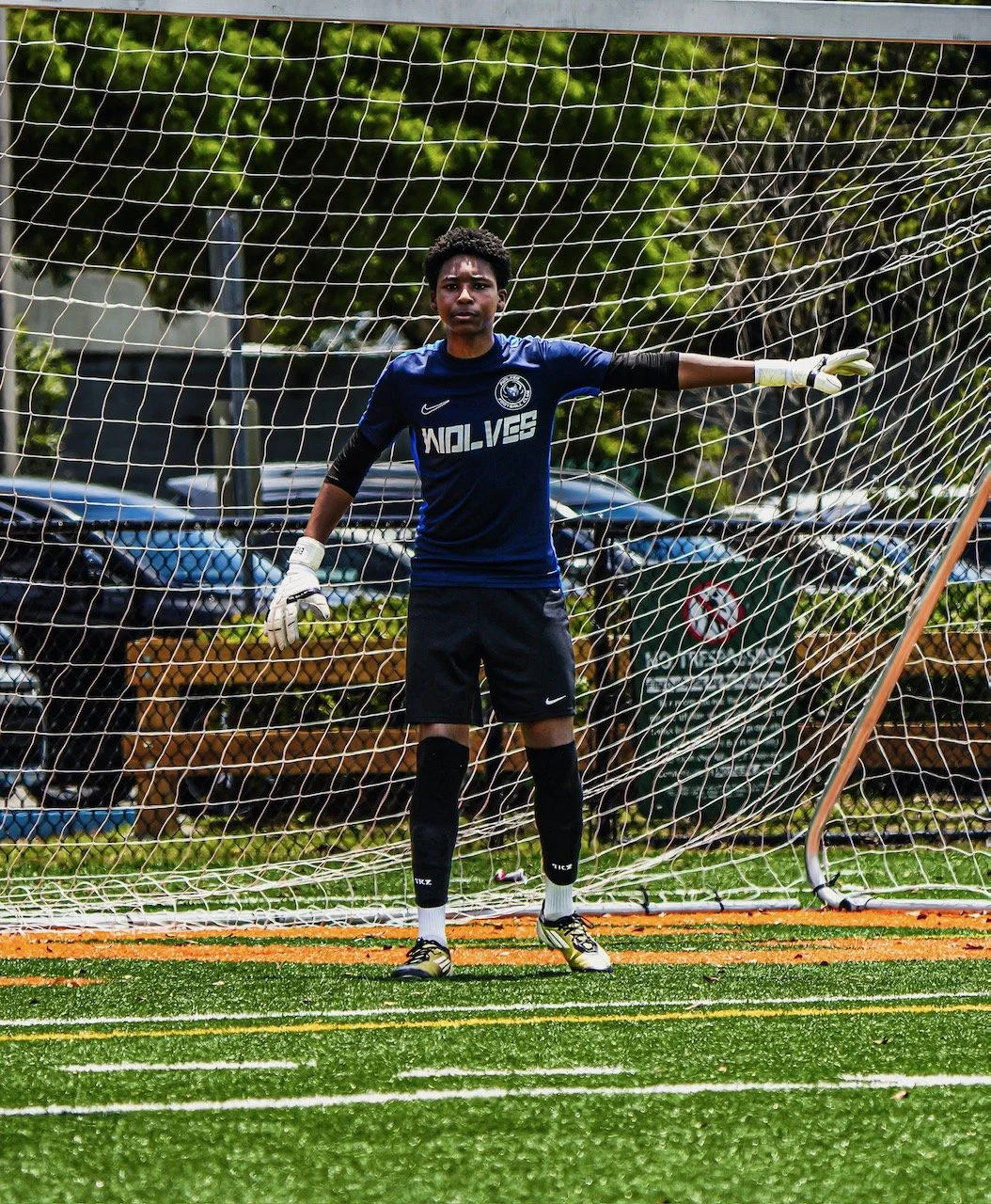 A young male soccer goalkeeper standing in front of the goal on a soccer field, wearing a blue uniform with 'WOLVES' written on it, gloves, and sports shoes.