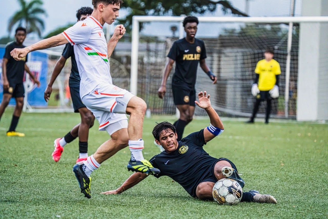 Soccer players in action, one sliding on the grass to kick the ball, others watching, with a goalkeeper in yellow in the background.
