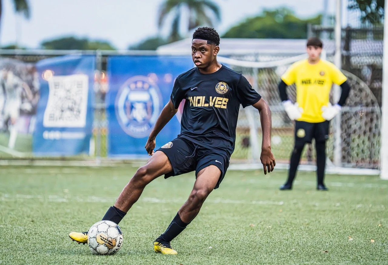 Young male soccer player in black uniform dribbling a soccer ball on the field, with goalkeeper in yellow jersey standing behind him.