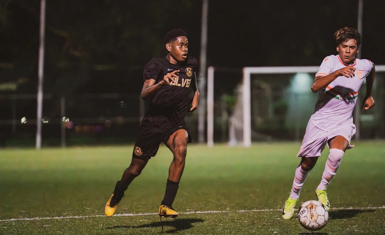 Two young male soccer players in action on a soccer field at night. One player in a black uniform is running, while the other in a white uniform is dribbling the ball.
