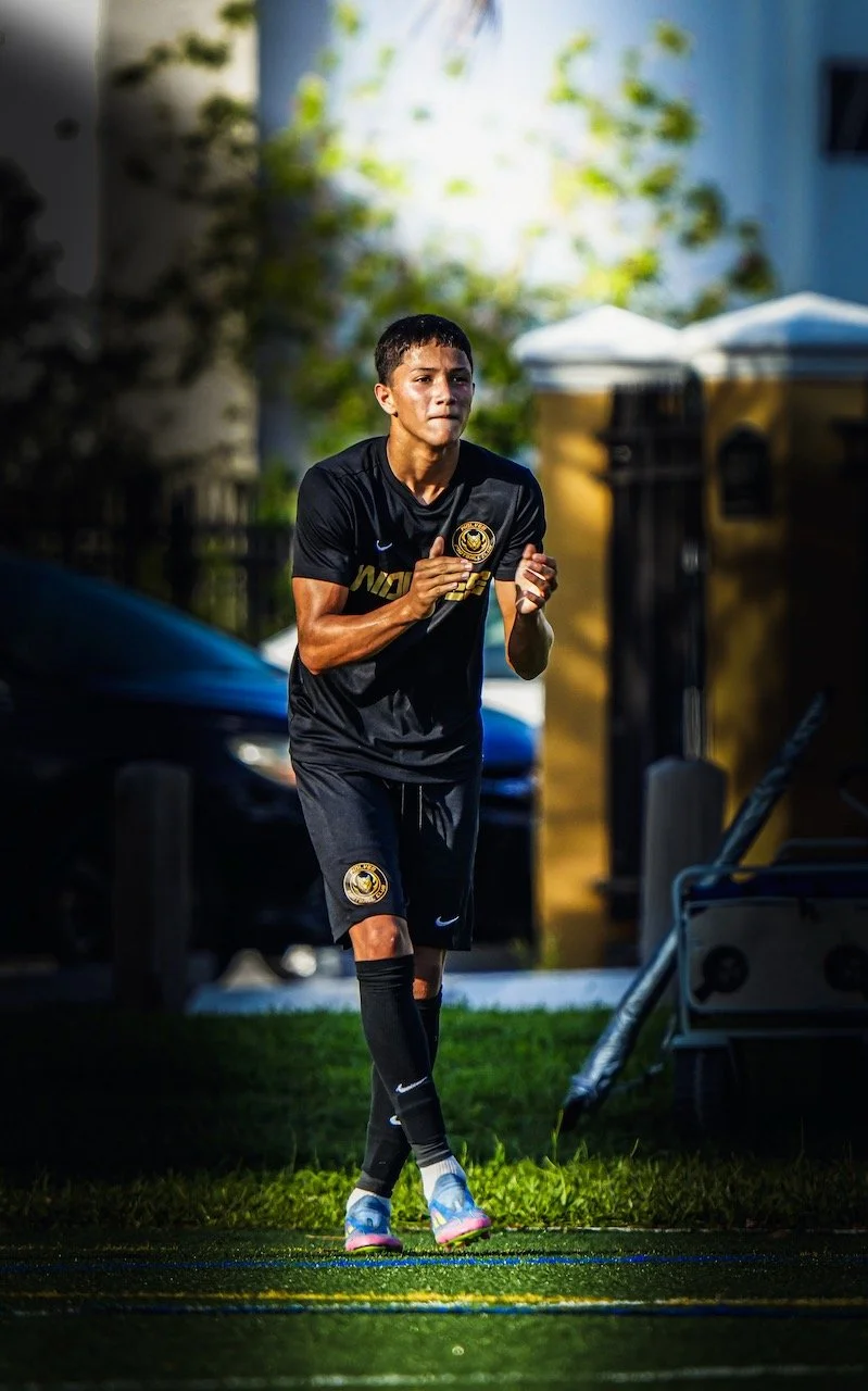 A young male soccer player in a black uniform jogging on a field during daytime.