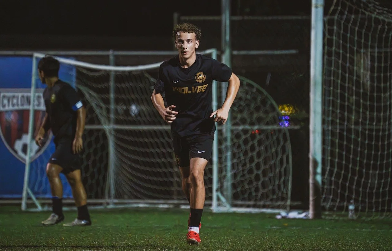 A young male soccer player running on the field at night during practice or game, wearing a black uniform with gold logo and text, with goalpost and net in the background.