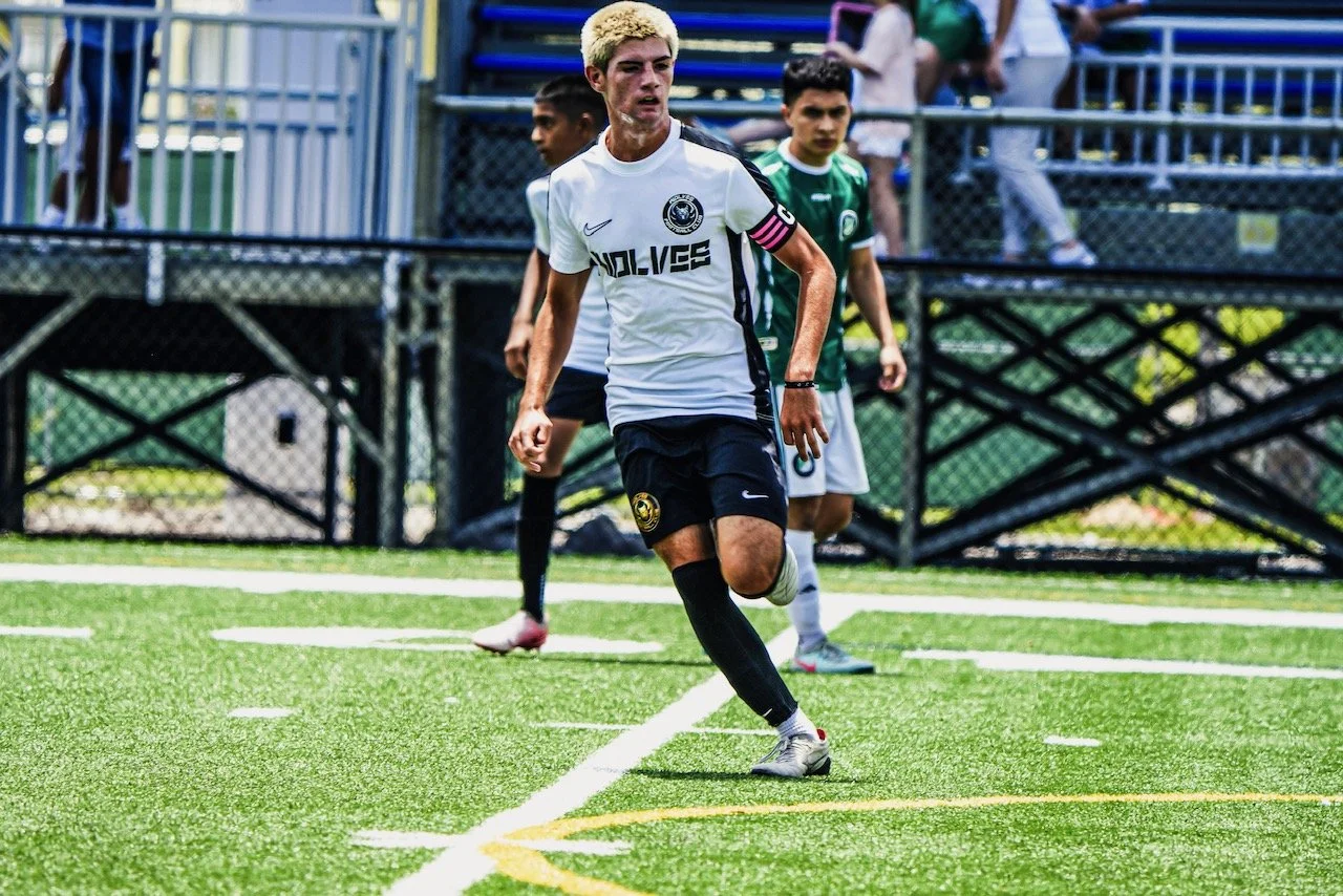 A soccer player with blond hair in a white jersey with black shorts and black socks is running on a green field during a match. There are other players and spectators in the background.