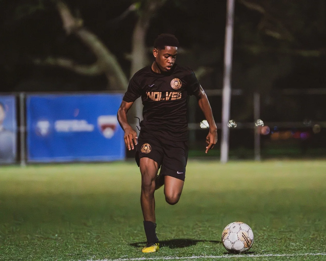 Young male soccer player in black uniform practicing on a field at night, kicking a soccer ball.