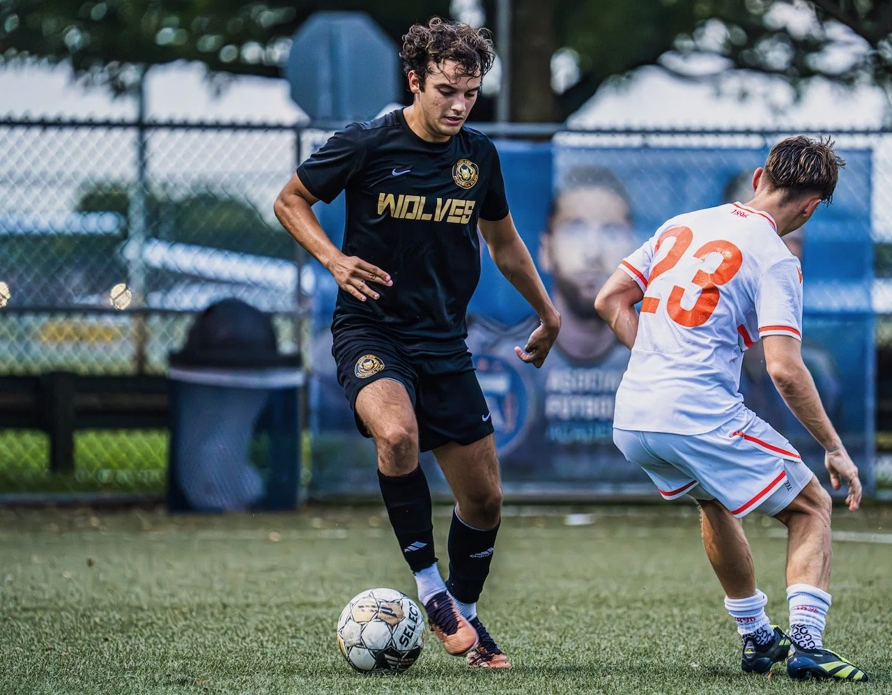 Two soccer players compete for the ball on a field. The player on the left is wearing a black jersey with 'WOLVES' written on it and black shorts, while the player on the right is dressed in a white jersey with orange accents and the number 23, and w