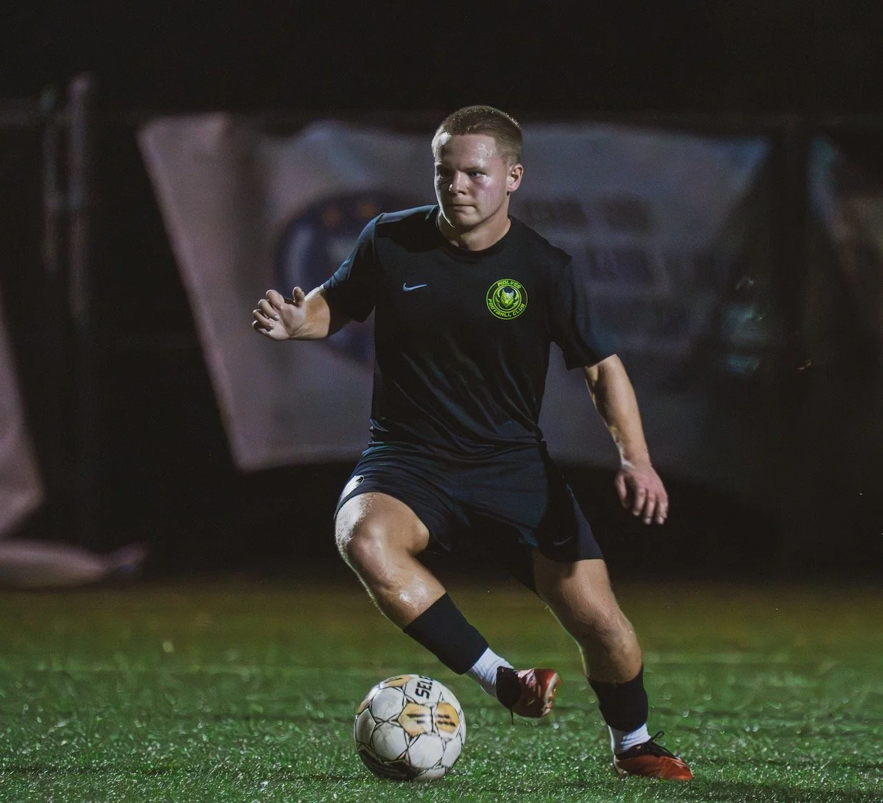 A male soccer player wearing a black jersey and shorts, kicking a soccer ball on a green field at night.