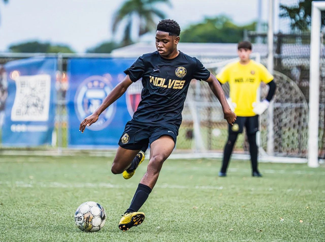 A young male soccer player wearing a black jersey and shorts is kicking a soccer ball on a grassy field during daytime. There is a goalkeeper in a yellow jersey standing near the goal in the background.