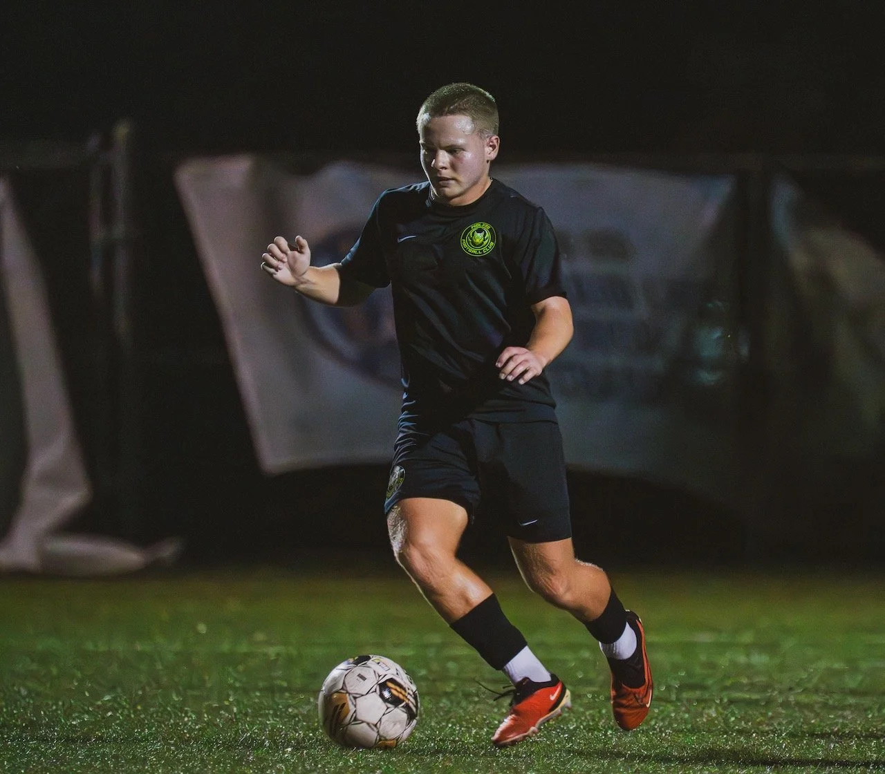 A male soccer player with blonde hair in black uniform with a green and yellow emblem, running on a grass field during night, controlling a soccer ball.