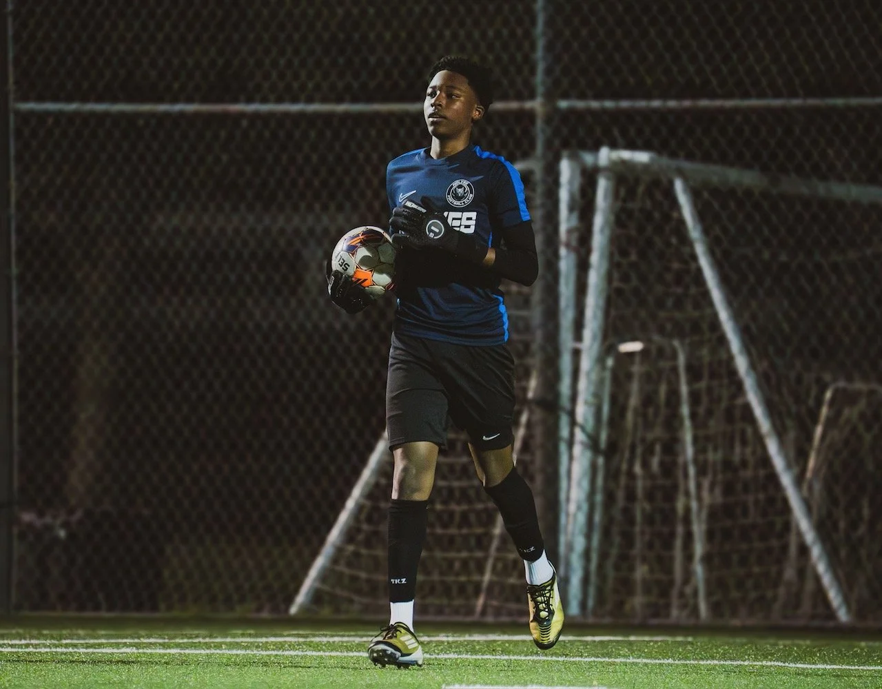 A young male soccer player in a blue jersey, black shorts, and yellow cleats running on a soccer field at night while holding a soccer ball and wearing goalkeeper gloves.
