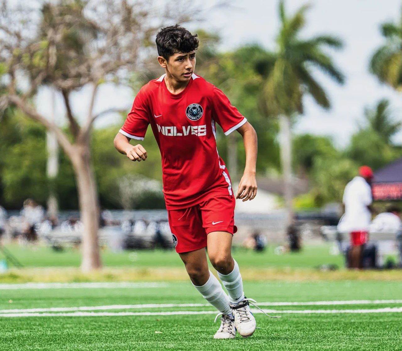 A young male soccer player in a red jersey and white socks running on a soccer field during daytime.