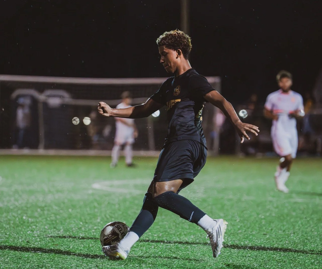 A young soccer player in a black uniform kicking a ball on a green field at night, with other players and a goal in the background.