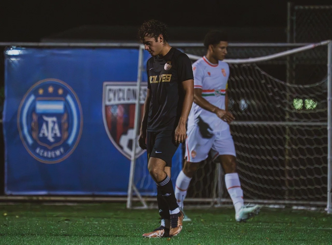 Two soccer players wear black and white uniforms on a field near a goal during a nighttime game.