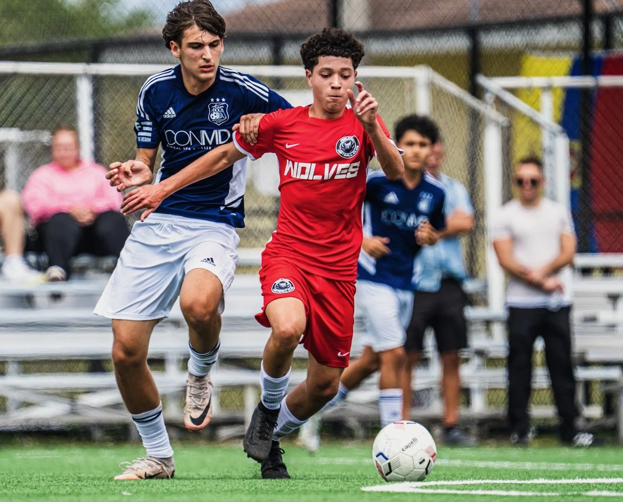 Two young soccer players compete for the ball, one in a blue jersey and the other in a red jersey, on a grassy field during a match.