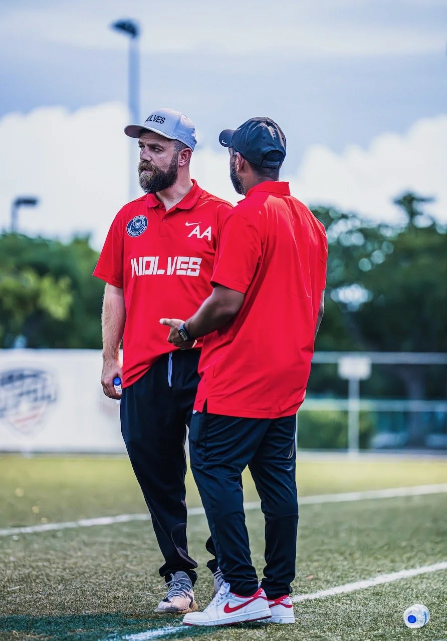 Two men in red shirts and black pants standing on a sports field, engaging in conversation, with trees and a cloudy sky in the background.