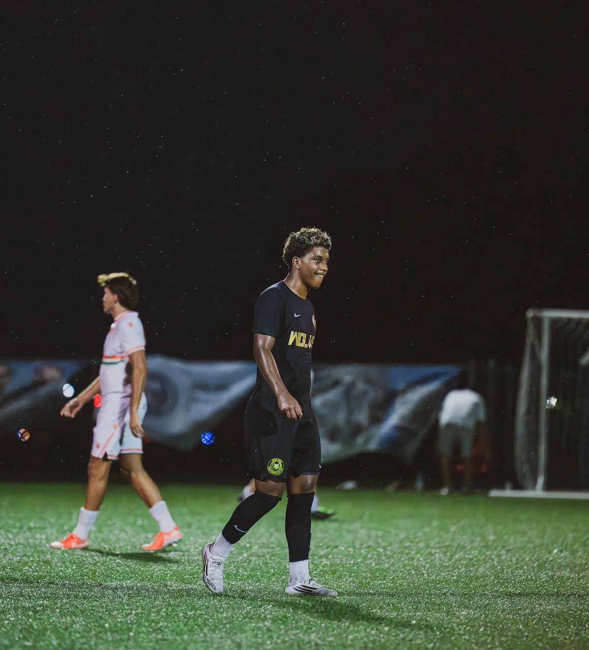 A female soccer player standing on the field at night with a slight smile, wearing a black uniform, with another player in a white uniform blurred in the background.