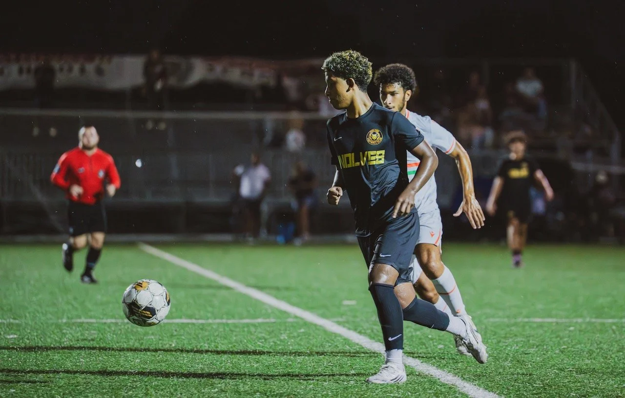 Two soccer players competing for the ball on a field during a night match, with a referee running in the background and spectators watching from the stands.
