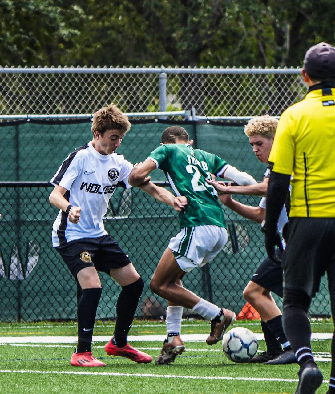 Young soccer players competing for the ball on a field, with a referee observing, in an outdoor setting with a chain-link fence and trees in the background.