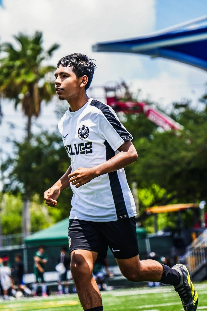 Young male soccer player running on a field, wearing a white jersey with black accents and black shorts, with trees and playground equipment in the background.