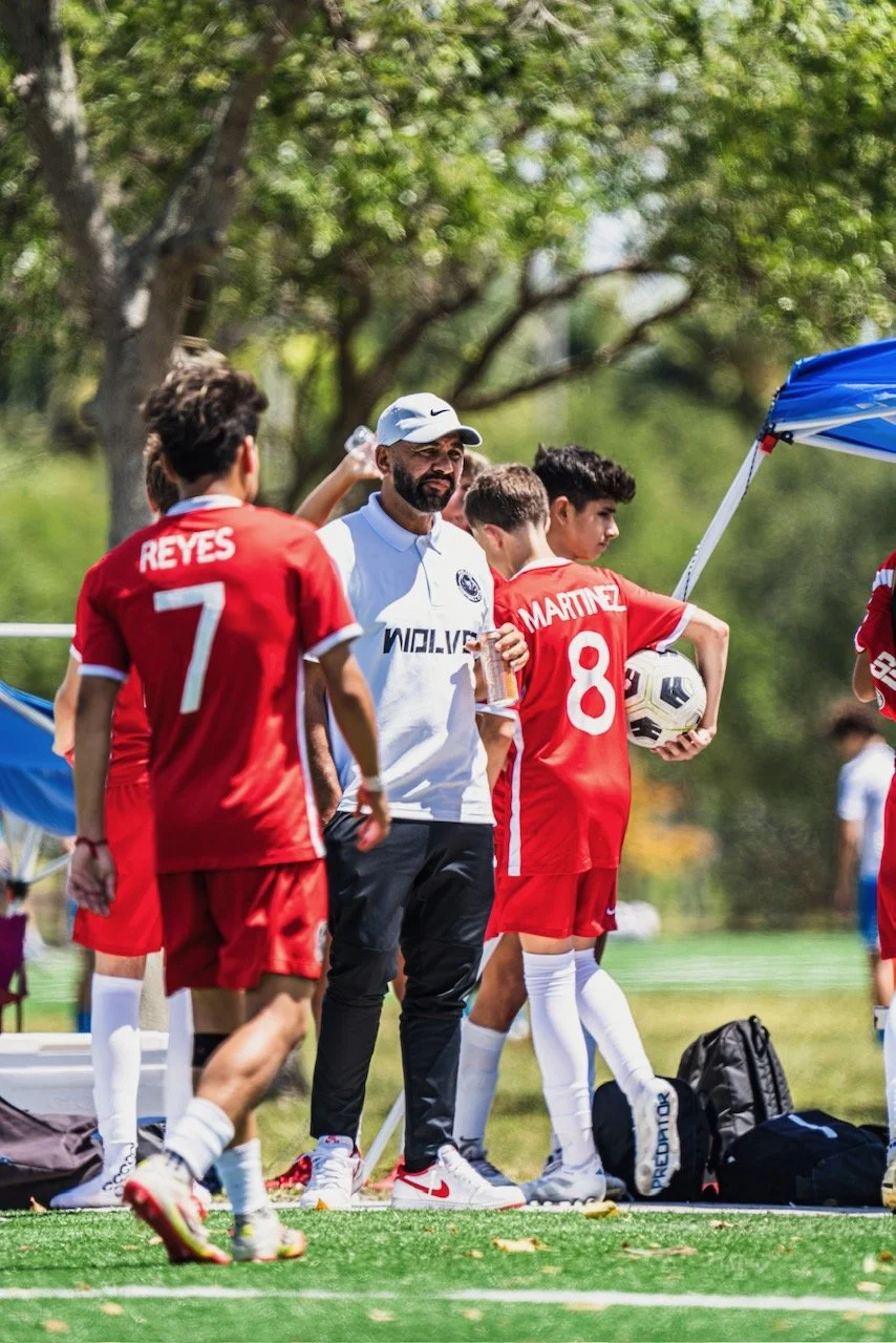 A group of young soccer players in red jerseys gathered around their coach, who is holding a water bottle and talking to them, on a soccer field with trees in the background.