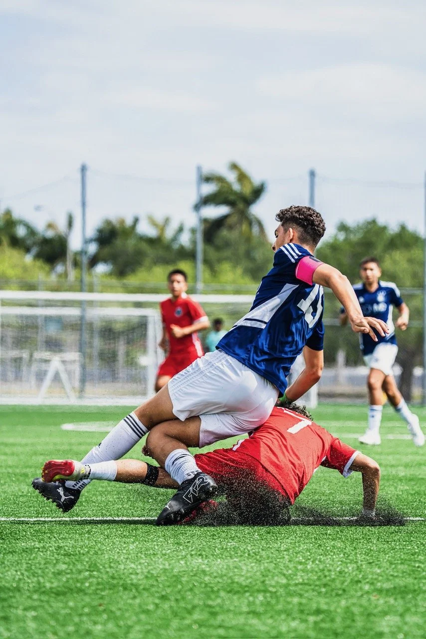 Soccer match showing a player in a blue jersey falling over a player in a red jersey on the field, with other players in the background.