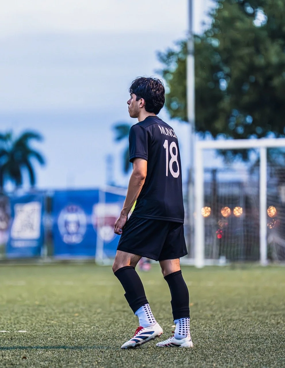 A male soccer player standing on a field, wearing a black jersey with the number 18 and the name MUNOZ on the back, black shorts, and white soccer cleats with black and red accents.