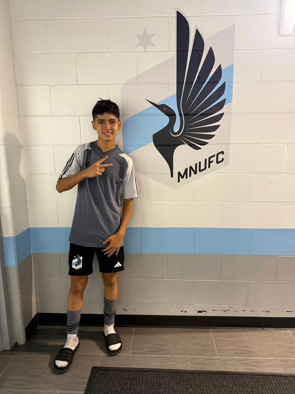Young boy in sportswear standing next to a Manchester United logo on a wall, making a peace sign with his right hand.
