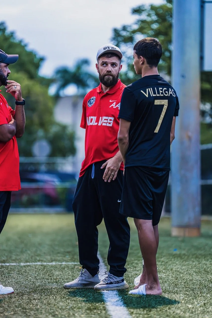 Three men standing on a field near a pole, having a conversation. The man in the center has a beard, wears a red sports shirt with logos, a gray cap, and black pants. The man on the right has short dark hair, wears a black sports shirt with the name