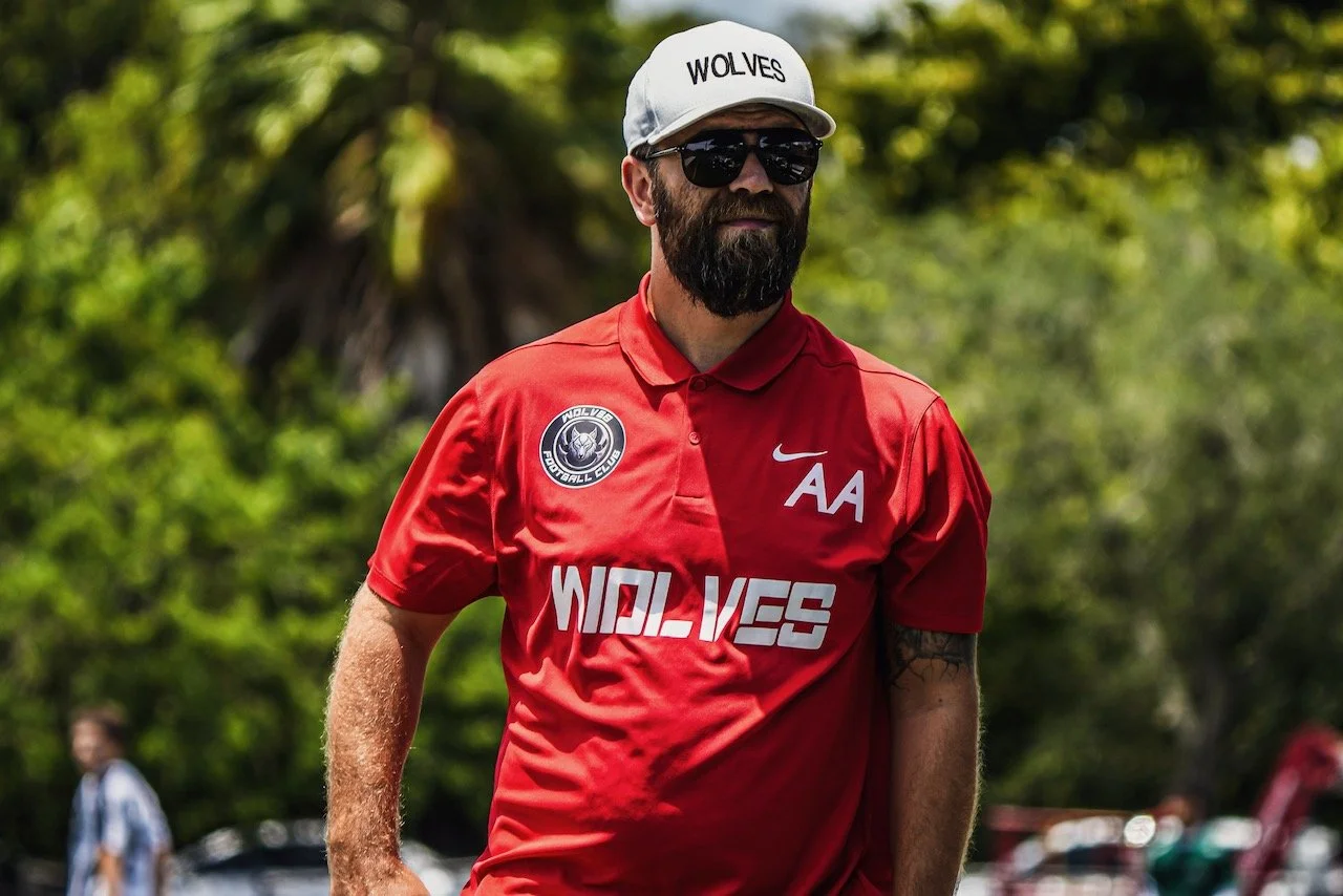 A man wearing a red sports shirt and a gray cap with the word 'WOLVES' stands outdoors, wearing sunglasses with a background of green trees.