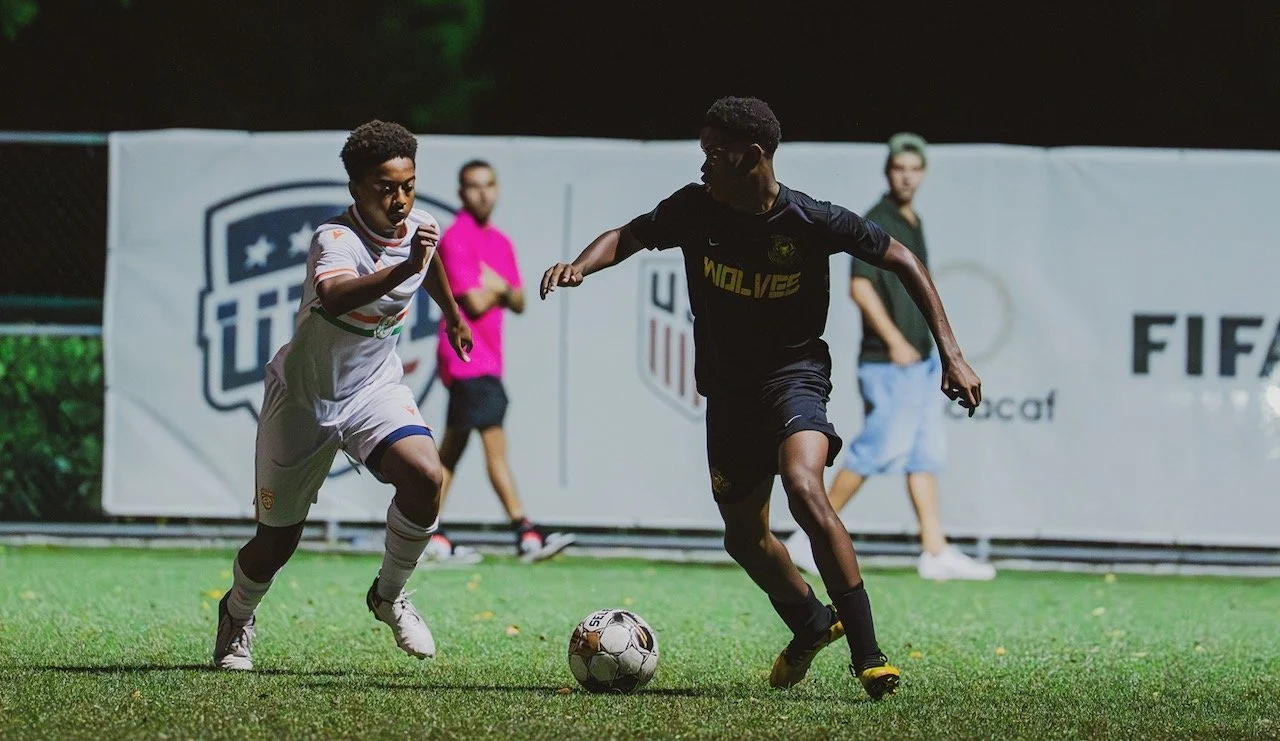 Two young boys playing soccer on a field at night, with one wearing a white jersey and the other in a black jersey, with FIFA banners in the background.