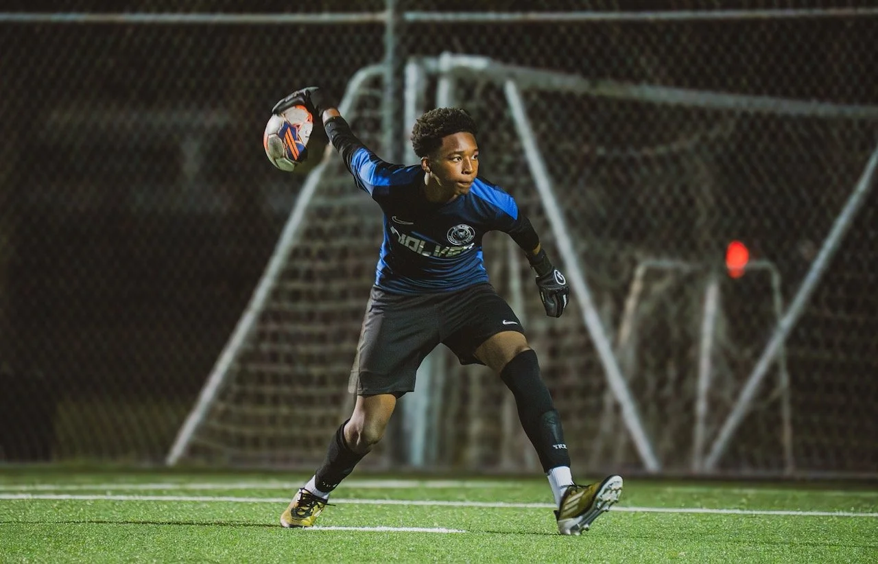 A young goalkeeper in black and blue uniform making a save during a night soccer game, with a goal net in the background.