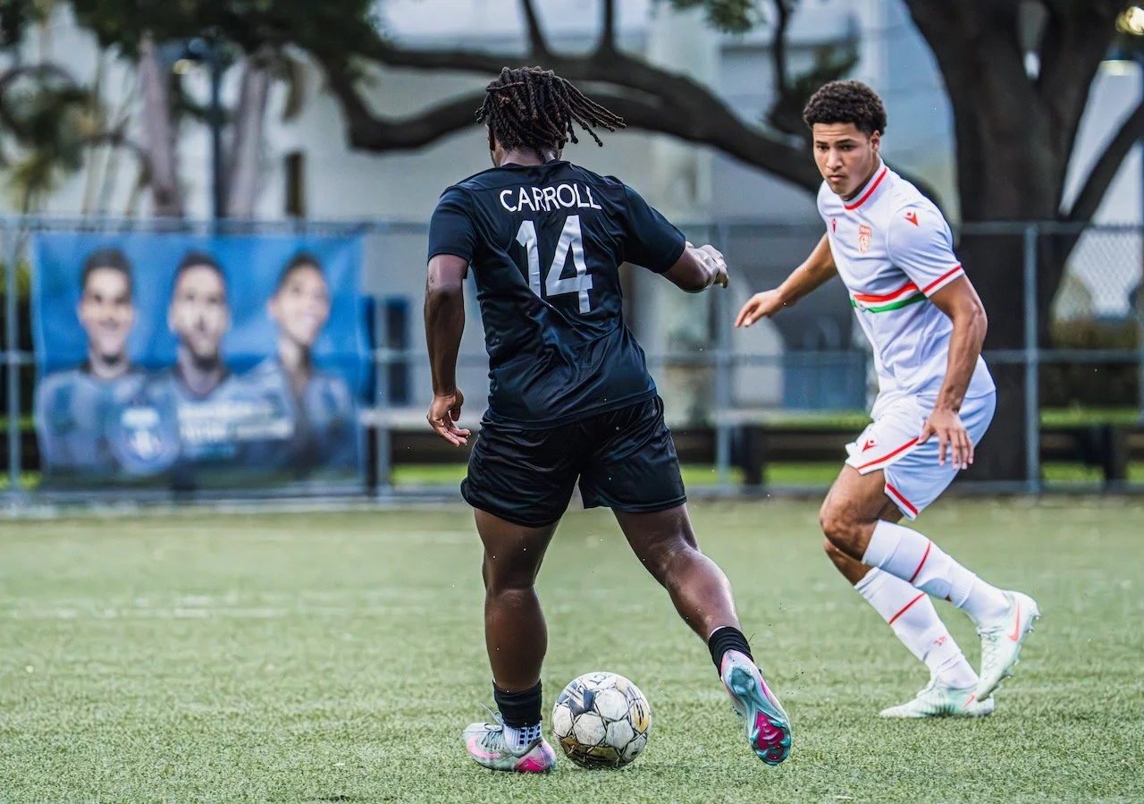Two young men playing soccer on a field, one wearing a black jersey with the name Carroll and the number 14, and the other in a white jersey with red and green stripes. The player in black is about to kick the ball while the player in white eyes him,