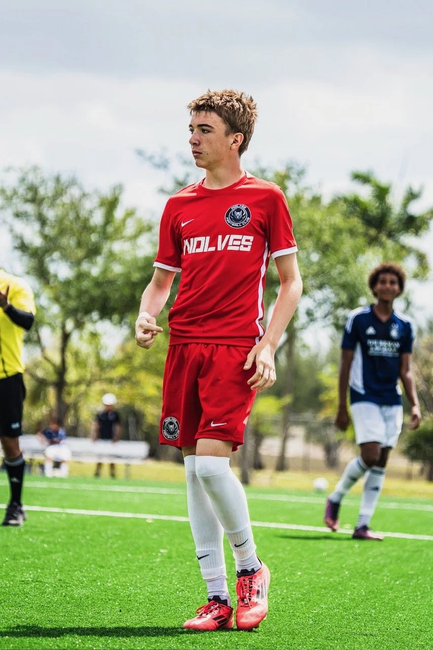 A young male soccer player wearing a red jersey and white socks on a soccer field, with other players in the background, during daytime with cloudy skies.