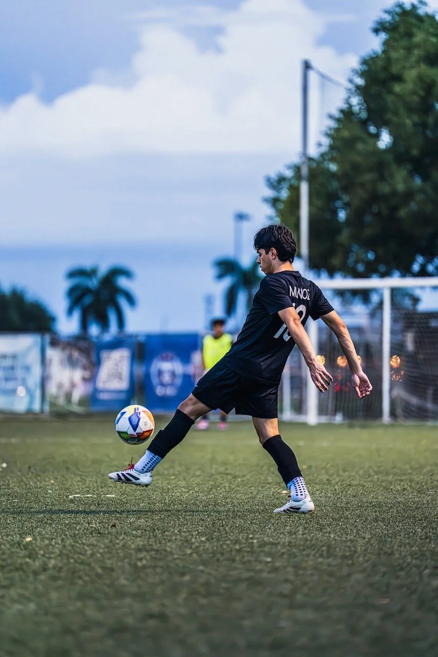 A soccer player in black uniform kicking a ball on a field during dusk, with palm trees and a goalpost in the background.