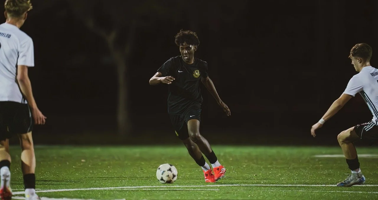 A soccer player in black uniform dribbling a soccer ball during a game at night with two opposing players nearby on a grassy field.