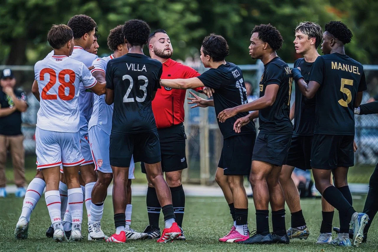 Two soccer teams, one in white and the other in black, are involved in a heated argument on the field, with players and a referee in the center.