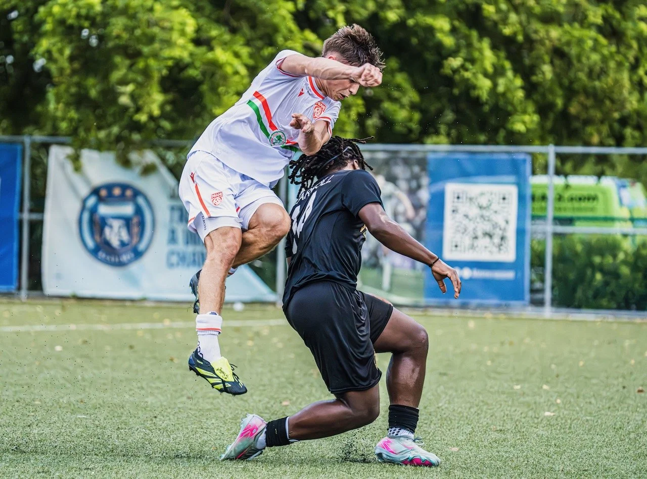 Two male soccer players competing for the ball on a grassy field, with trees and advertising banners in the background.